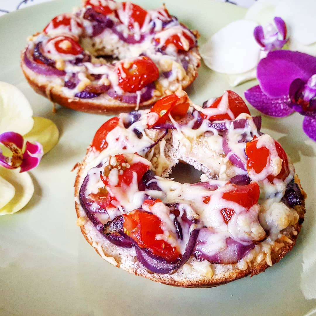 Two cheesy baked bagels topped with cherry tomatoes and red onions on a green plate, surrounded by purple and white orchids.