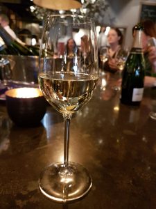 A close-up of a wine glass filled with white wine, set on a bar counter with a blurred background of people enjoying drinks.