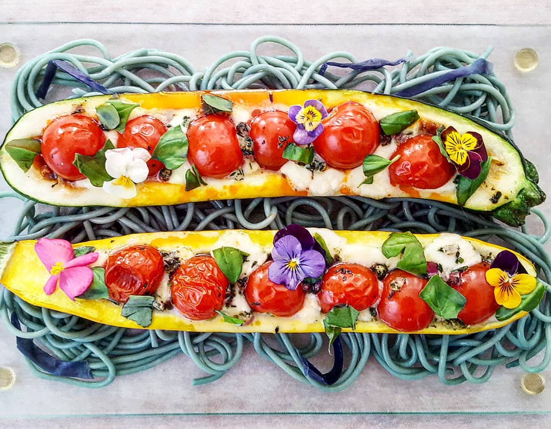 Two stuffed courgettes topped with cherry tomatoes, herbs, and edible flowers, served on a bed of blue pasta.